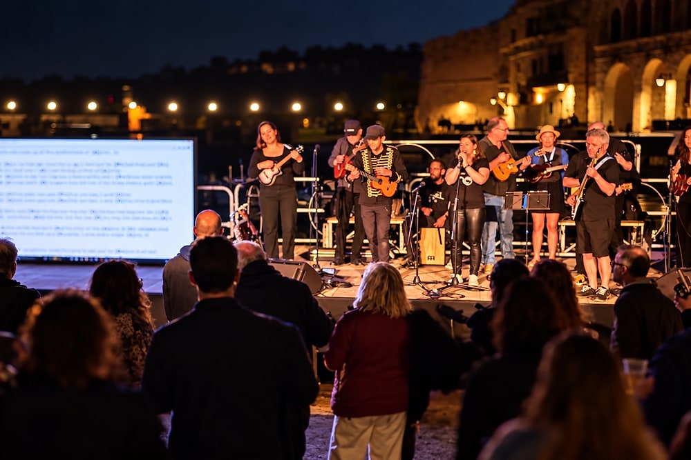 Ukulele Tuesday leading a play-along on the port of Monopoli at night: the band on stage, the chords on a big screen and a crowd playing along