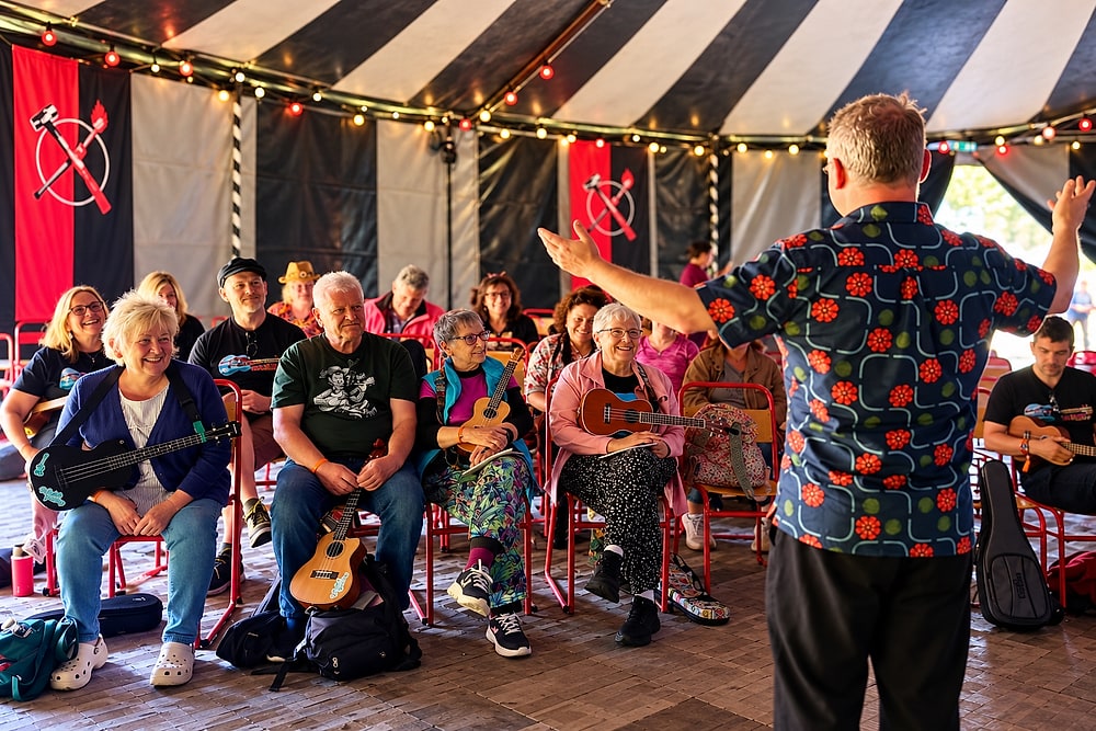 One of our members leading a songwriting workshop in a striped festival tent, arms wide, in front of a seated group holding ukuleles